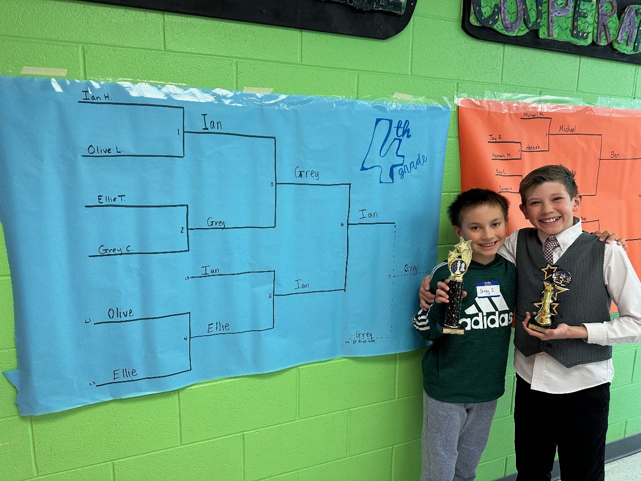 Two students holding trophies by a tournament bracket.